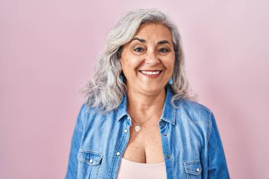 Yüzünde mutlu ve havalı bir gülümsemeyle pembe arka planda duran gri saçlı orta yaşlı bir kadın. şanslı kişi. 
