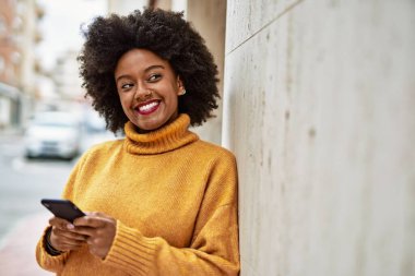 Genç Afrikalı Amerikalı kız şehirde akıllı telefon kullanarak mutlu bir şekilde gülümsüyor..