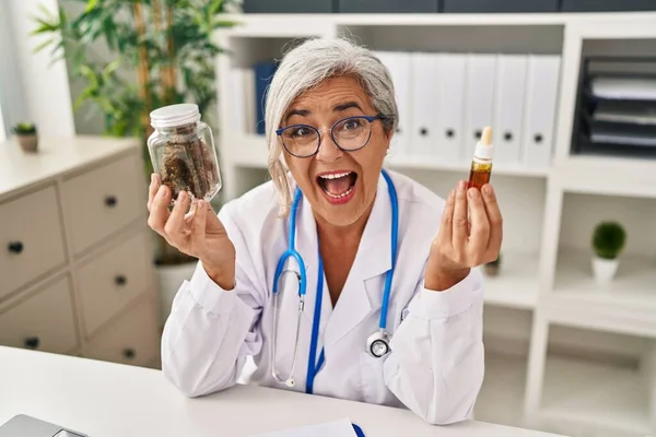 Middle age woman doctor holding cbd oil smiling and laughing hard out loud because funny crazy joke.