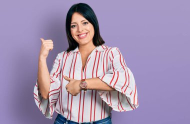 Gündelik giysiler giyen genç İspanyol kız arka tarafı işaret ediyor. Elleri havada, kendinden emin gülümsüyor. 