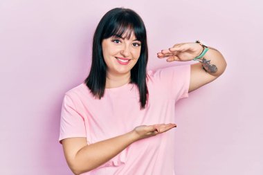 Latin kökenli genç bir kadın, gündelik kıyafetler giyiyor. Elleriyle işaret ediyor. Ölçü sembolü. Kameraya bakıp gülümsüyor. ölçüm kavramı. 