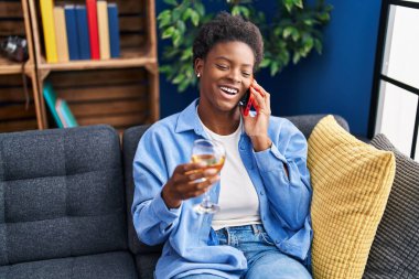 Afro-Amerikalı kadın akıllı telefondan konuşuyor evde şampanya içiyor.