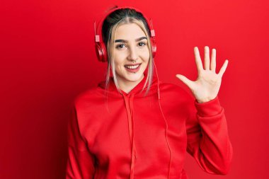 Genç, modern bir kız spor kıyafetleri giyiyor ve kulaklık takıyor. Beş numarayı gösterip işaret ediyor. Kendine güvenen ve mutlu bir şekilde gülümsüyor.. 