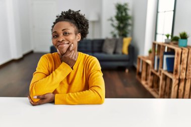 Genç Afro-Amerikan kadın, gündelik giysiler giymiş evde masada oturmuş kameraya bakıyor kollarını kavuşturup gülümsüyor ve çenesini kaldırıyor. Olumlu düşünmek. 