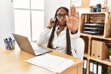 Ofiste örgü ören siyahi kadın telefonla konuşuyor. Avuç içiyle şarkı söylemeyi kes. Yüzünde olumsuz ve ciddi bir jest olan uyarı ifadesi. 