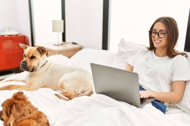 Genç İspanyol kadın, yatak odasında köpeklerle birlikte dizüstü bilgisayar kullanıyor.