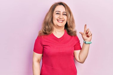 Orta yaşlı beyaz kadın, günlük kıyafetler giymiş, yüzünde kocaman bir gülümseme var. Elinle kameraya bakıyor.. 