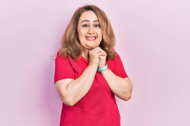 Orta yaşlı beyaz kadın, sıradan kıyafetler giyiyor. Gergin ve heyecanlı, elleri çenesinde, yan tarafa bakıyor. 