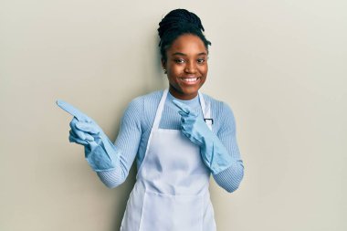 Saçları örgülü, daha temiz önlüklü, eldivenli, gülümseyen ve kameraya bakan Afro-Amerikalı bir kadın.. 