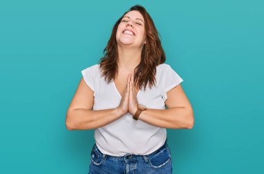 Genç ve cüsseli bir kadın günlük beyaz t-shirt giyip elleri ile yalvarıyor ve dua ediyor. Yüzünde umut ifadesi var. Çok duygusal ve endişeli. Yalvarıyor. 