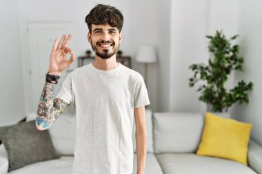 Oturma odasında sakallı İspanyol bir adam gülümsüyor ve el ve parmaklarıyla 