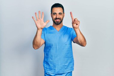 Sakallı, mavi erkek hemşire üniforması giyen yakışıklı İspanyol bir adam. Altıncı parmaklarını gösterip gösterirken kendinden emin ve mutlu gülümsüyor.. 