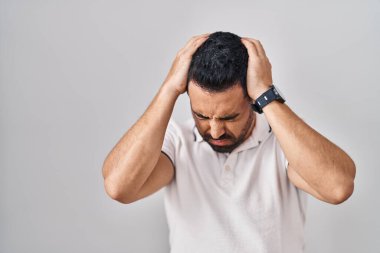 Beyaz arka plan üzerine günlük kıyafetler giyen sakallı genç İspanyol adam acı ve migren yüzünden çaresiz ve stresli bir baş ağrısı çekiyor. Eller başının üstünde.. 