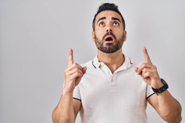 Beyaz arka plan üzerine günlük kıyafetler giyen genç İspanyol adam şaşırmış ve şaşırmıştı. Parmaklarıyla işaret ediyor ve kollarını kaldırıyordu.. 