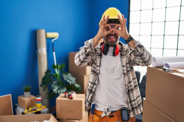 Sakallı, evde tadilat yapan genç bir İspanyol. Dürbün gibi jest yapıyor. Gözleri parmaklarından bakıyor. çılgın ifade. 