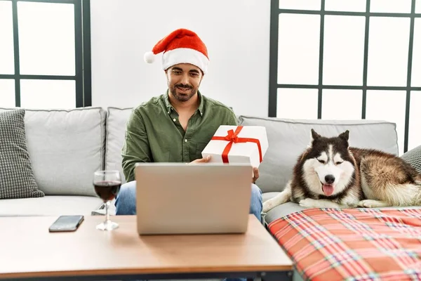 Young hispanic man having video call holding gift and sitting on sofa with dog at home