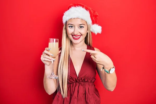 Молодая Блондинка Рождественской Шляпе Пьет Шампанское Улыбаясь Показывая Пальцем Рукой — стоковое фото
