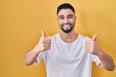 Sarı arka plan işareti üzerine sıradan bir tişört giyen genç yakışıklı. Elleriyle olumlu jest yapıyor. Gülümsüyor ve mutlu. Neşeli ifade ve kazanan jest. 