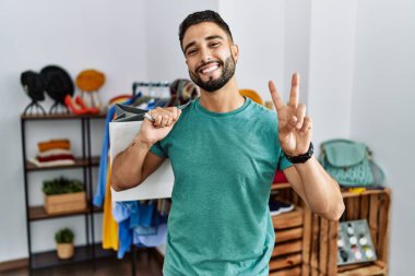 Satış mağazasında sakallı, alışveriş torbası tutan genç yakışıklı adam kameraya bakıp zafer işareti yapıyor. İki numara.. 