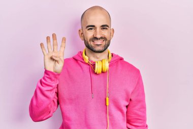 Spor kıyafetleri giyen ve kulaklık takan genç kel bir adam dört numarayı gösterip işaret ederken kendinden emin ve mutlu bir şekilde gülümsüyor.. 