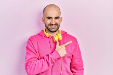 Spor kıyafetleri giyen ve kulaklık takan genç kel bir adam. Yüzünde mutlu ve doğal bir ifadeyle, elleri ve parmaklarıyla gülümseyen bir yüz ifadesi var. 