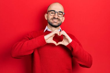 Gündelik giysiler ve gözlük takan genç kel bir adam aşkta gülümsüyor. Elleriyle kalp sembolü yapıyor. romantik kavram. 