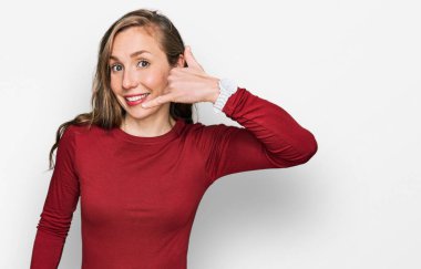 Sıradan kıyafetler giyen, gülümseyen, el ve parmaklarıyla telefon hareketi yapan, telefonda konuşan genç sarışın bir kadın. iletişim kavramları. 