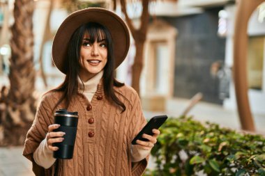 Kışlık şapkalı esmer kadın akıllı telefon kullanıyor ve şehirde bir fincan kahve içiyor.