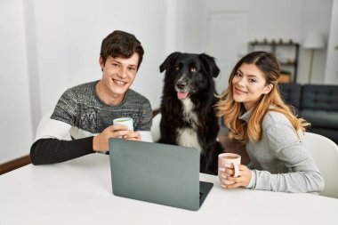 Genç beyaz çift dizüstü bilgisayar kullanıyor ve kahve içiyor. Evde köpeklerle birlikte masada oturuyorlar..