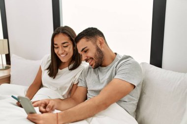 Genç Latin çift evde yatarken akıllı telefon kullanıyor..