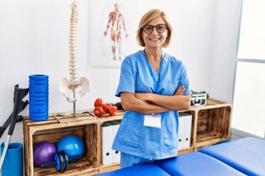 Orta yaşlı, sarışın, fizyoterapi üniforması giyen bir kadın klinikte kollarını kavuşturmuş duruyor.