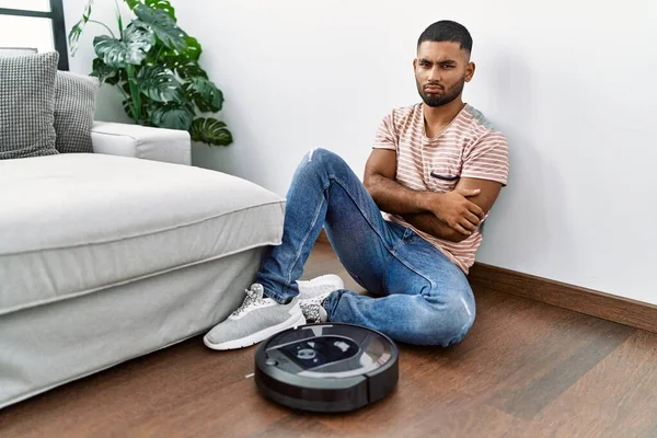 Young indian man sitting at home by vacuum robot skeptic and nervous, disapproving expression on face with crossed arms. negative person.