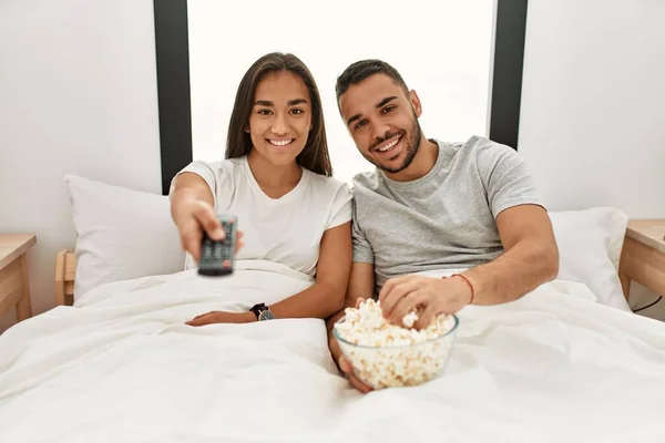 Young latin couple watching movie and eating popcorn lying on bed.