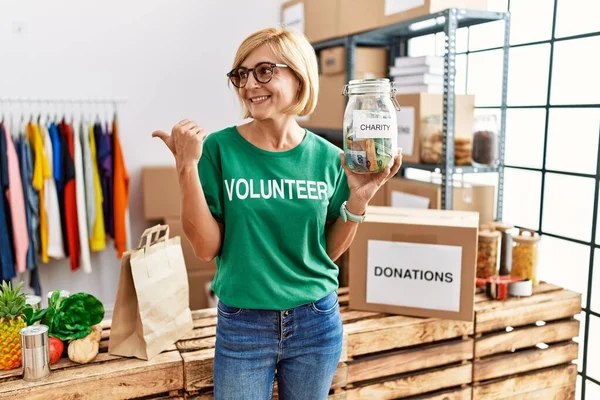 Orta yaşlı, sarışın, gönüllü tişört giyen, yardım parası tutan, ağzı açık gülümseyen bir kadın. 
