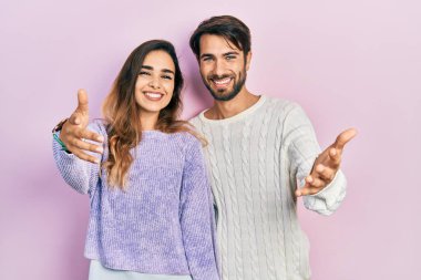 Gündelik giysiler giymiş genç İspanyol bir çift kameraya bakıp sarılmak için kollarını açmış gülümsüyor. Mutluluğu kucaklayan neşeli bir ifade.. 