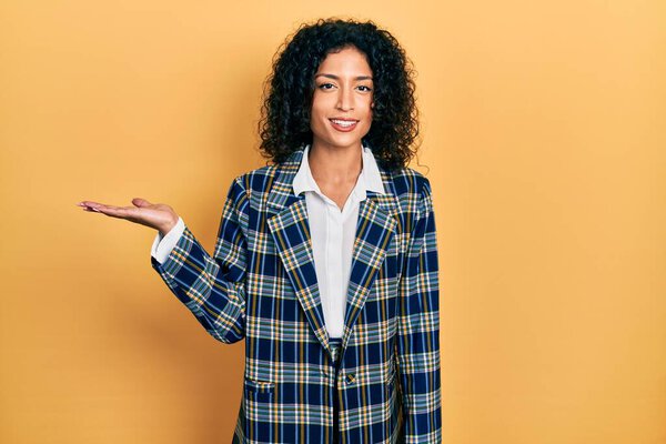 Молодая латиноамериканка в деловой одежде улыбается веселым подарком и указывает ладонью на камеру. 