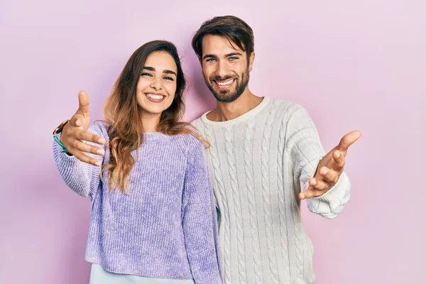 Gündelik giysiler giymiş genç İspanyol bir çift kameraya bakıp sarılmak için kollarını açmış gülümsüyor. Mutluluğu kucaklayan neşeli bir ifade.. 