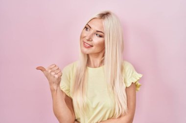 Pembe arka planda duran beyaz kadın gülümsüyor, gülümsüyor ve başparmağıyla yan tarafa bakıyor.. 