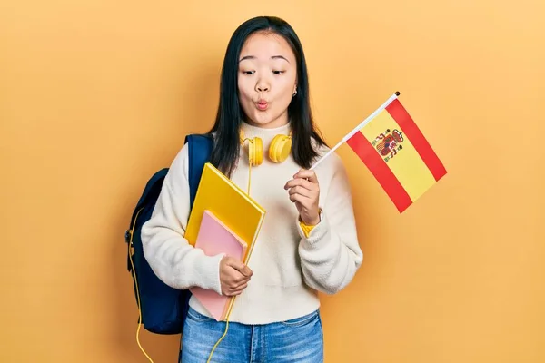 Young chinese girl exchange student holding spanish flag making fish face with mouth and squinting eyes, crazy and comical.