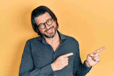 Orta yaşlı beyaz bir adam günlük kıyafetler ve gözlük takıyor gülümsüyor ve kameraya bakıyor iki eliyle ve parmaklarıyla yandan işaret ediyor.. 