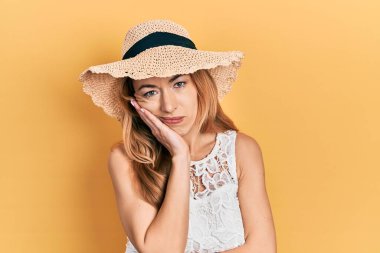 Yazlık şapka takan genç beyaz kadın kollarını kavuşturarak depresyondan sıkılmış ve yorgun görünüyor.. 