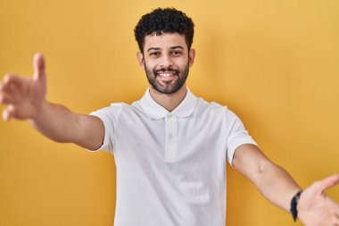 Arap adam sarı arka planda durup kameraya bakarak sarılmak için kollarını açmış gülümsüyor. Mutluluğu kucaklayan neşeli bir ifade.. 