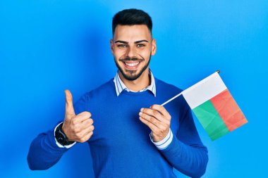 Sakallı genç İspanyol adam elinde madagaskar bayrağı, mutlu ve pozitif gülümsüyor, başparmağıyla mükemmel ve onay işareti yapıyor. 