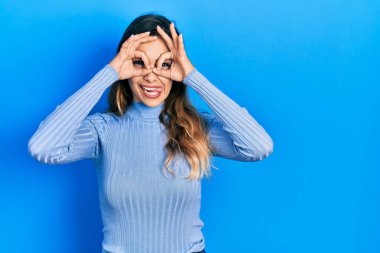 Günlük kıyafetler giyen genç İspanyol kız dürbün gibi jest yapıyor, gözleri parmaklarından bakıyor. çılgın ifade. 