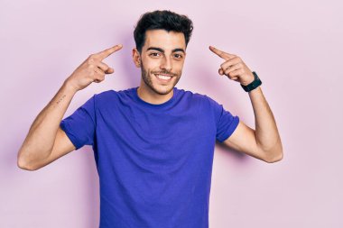 Gündelik tişört giyen genç İspanyol adam iki eliyle başını işaret ederek gülümsüyor, harika bir fikir ya da düşünce, iyi bir hafıza 