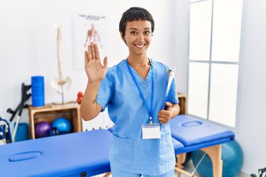 Kısa saçlı, İspanyol bir kadın ağrı kesici kliniğinde çalışıyor. Mutlu, güler yüzlü, dostça bir jest yapıyor. 