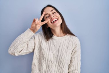 Young brunette woman standing over blue background doing peace symbol with fingers over face, smiling cheerful showing victory 