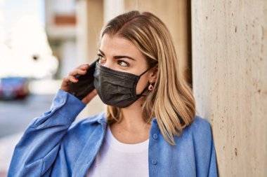 Coronavirus maskesi takan genç sarışın kız şehirdeki akıllı telefondan konuşuyor..