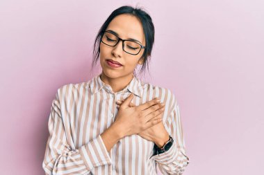 Gündelik kıyafetler ve gözlük takan genç İspanyol kız elleri göğsünde, gözleri kapalı gülümsüyor ve yüzünde minnet ifadesi var. sağlık kavramı. 