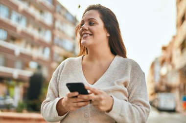 Genç İrlandalı artı cüsseli kız şehirde akıllı telefon kullanarak mutlu gülümsüyor..